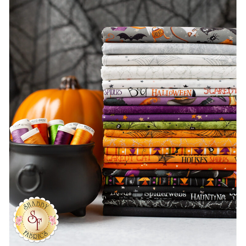 A stack of Halloween-themed fabric bolts in orange, black, purple, and gray colors is displayed beside a small black cauldron filled with colorful spools of thread. An orange pumpkin is placed in the background, against a web-like backdrop.