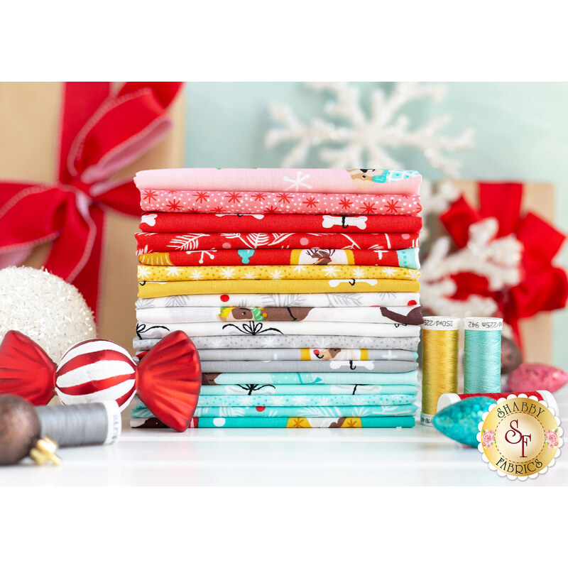 A stack of colorful fabric bundles sits on a white surface, surrounded by holiday decorations, including red bows, ornaments, and spools of thread. The fabrics feature various festive patterns in red, white, blue, and other colors.