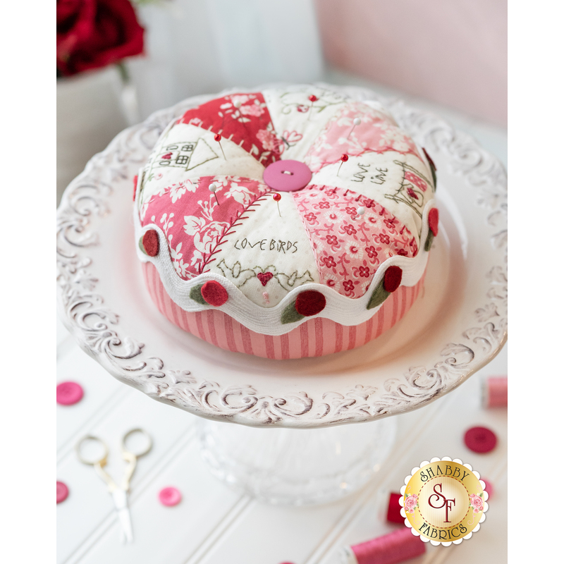 Love pinwheel pastry pin cushion on a decorated white cake stand with red roses in the background, all on a white tabletop with pink buttons and thread