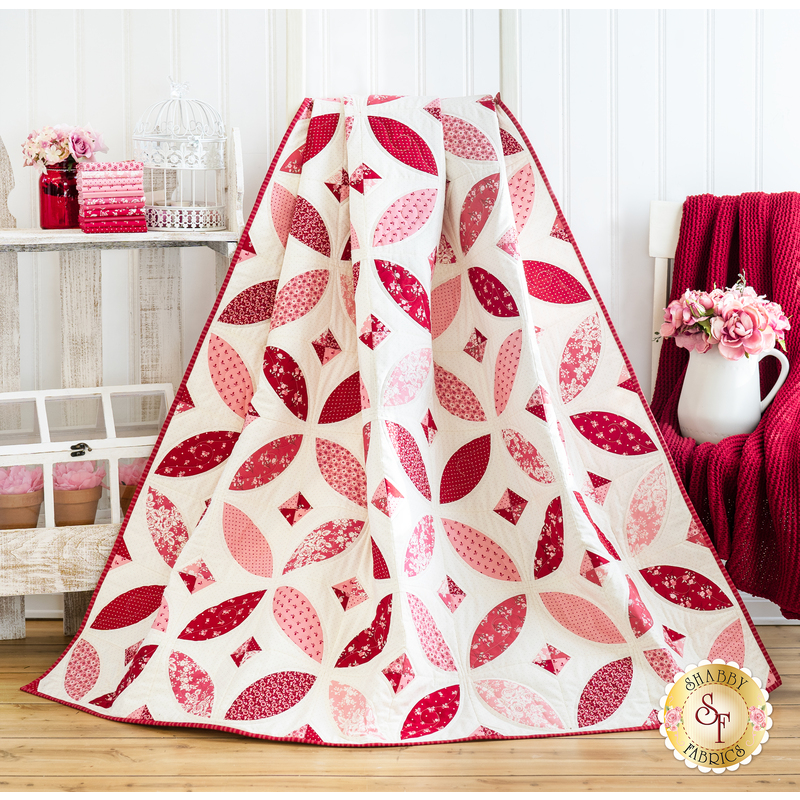 Completed quilt draped in front of a white wall with a chair draped with a red blanket and white shelf with decorations in the background