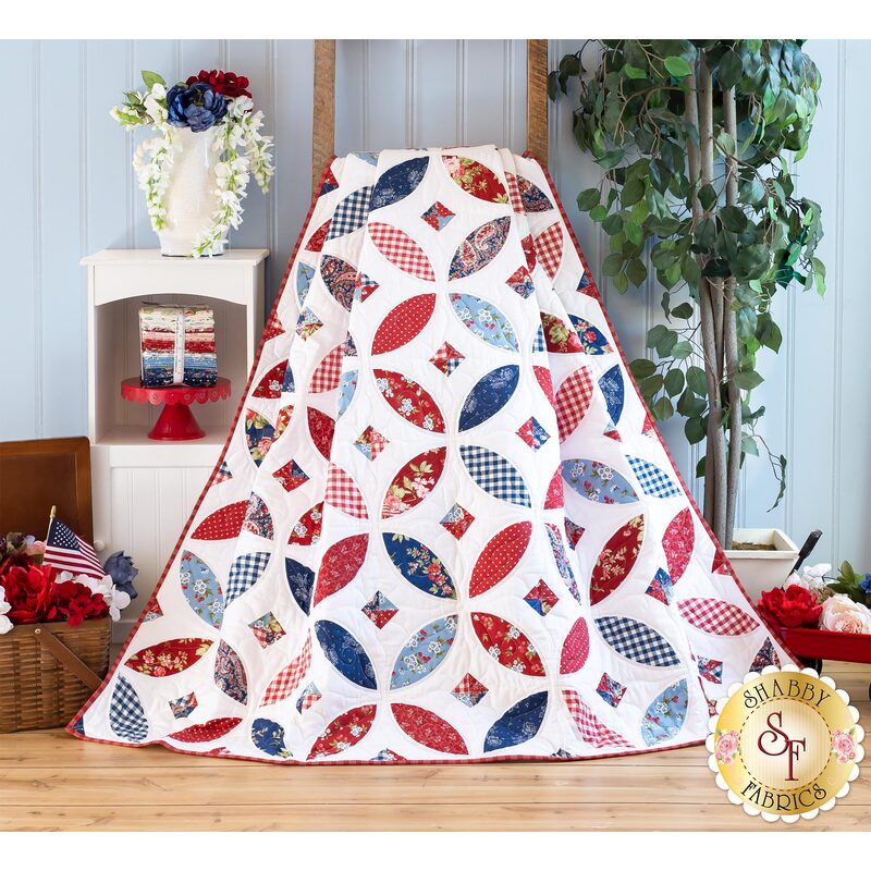 Photo of a red, white and blue quilt draped over a small brown ladder leaning against a light blue paneled wall with patriotic decor and a houseplant on either side.