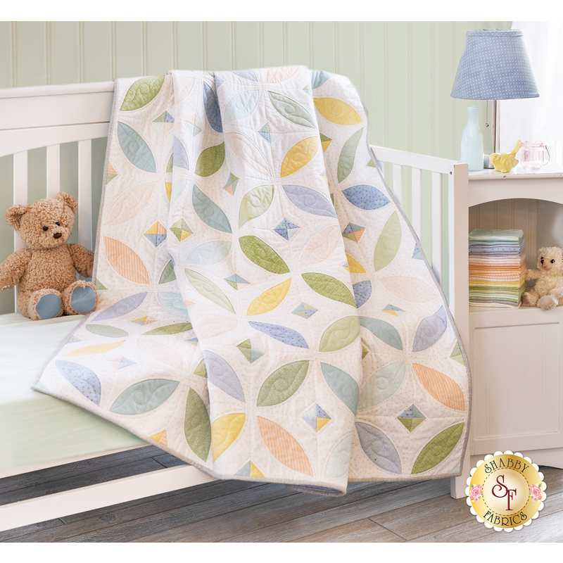 Orange Blossom Little Lambies Spring quilt draped over a white crib with a stuffed bear sitting next to it on the mattress and a shelf with a lamp and stack of fabric from the Little Lambies Flannel collection in the background