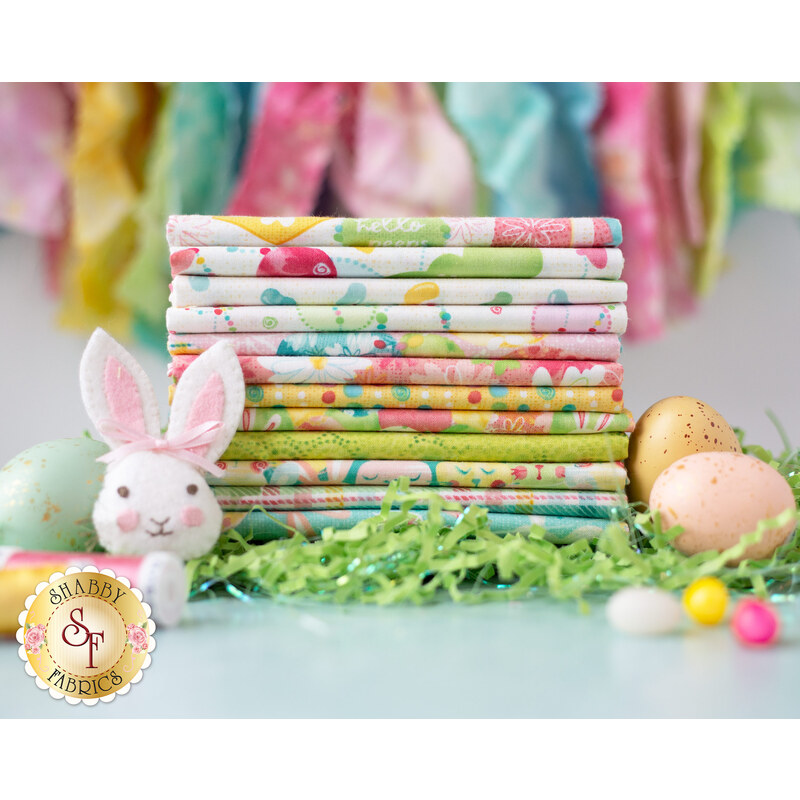 A stack of colorful fabrics with Easter-themed patterns sits on a table surrounded by pastel-colored Easter eggs and green grass-like filling. A small plush bunny with pink ears is in front of the fabric.