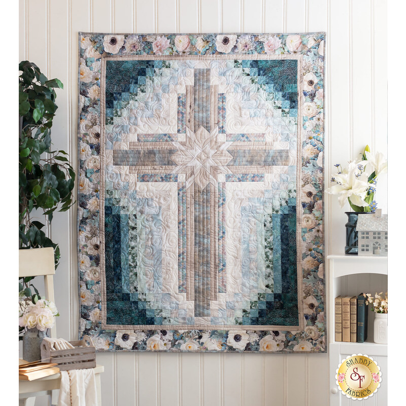 isolated image of a rectangular quilt that features a large cross motif in the center in muted floral fabrics with dark teal accents hanging on a white paneled wall with home decor items on a shelf and small chair with a house plant on either side of the quilt