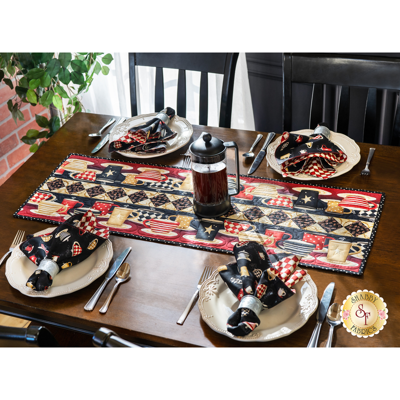 coffee-themed table runner with a checkered stripe and coffee mugs in neutral tones, red, and black on a brown dining table with a window and house plant in the background with a french press, and our place settings with matching napkins on the sides.