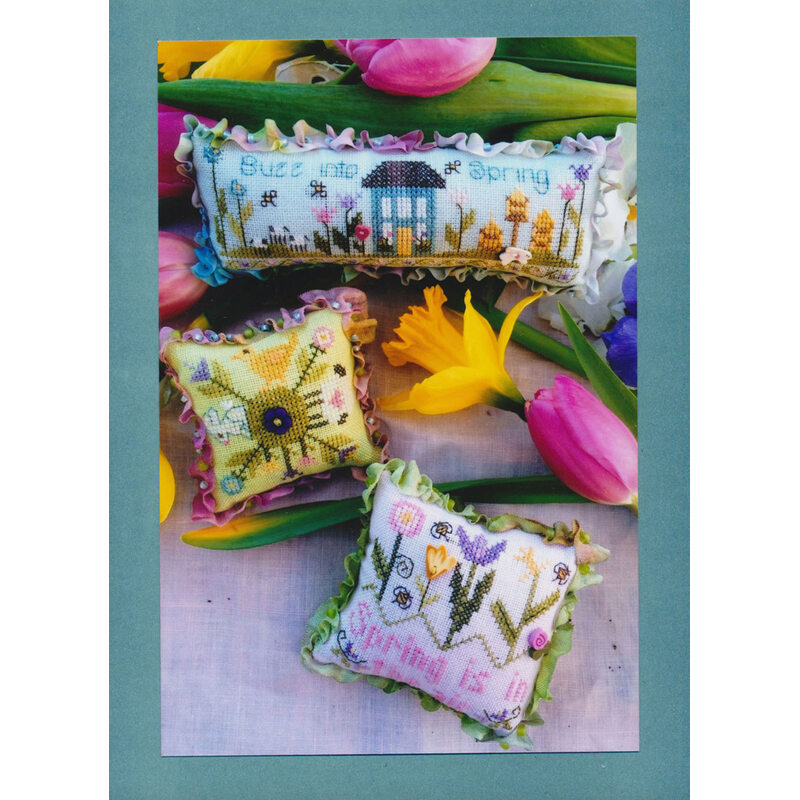 Three decorative cross stitch pillows with spring themes, surrounded by colorful tulips.