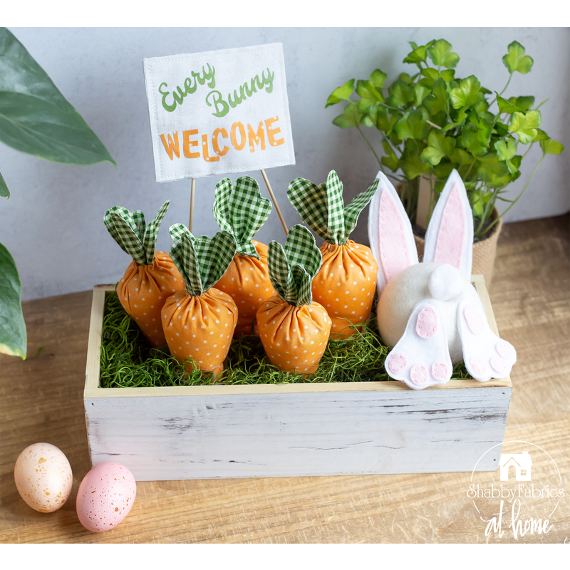 A white washed planter box filled with green moss and fabric carrots with a bunny figure diving into the box and a sign that says 