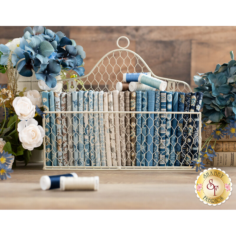 A wire basket filled with neatly arranged fabric bolts in various shades of blue, tan, and white, surrounded by blue and white flowers, with spools of thread in the foreground.