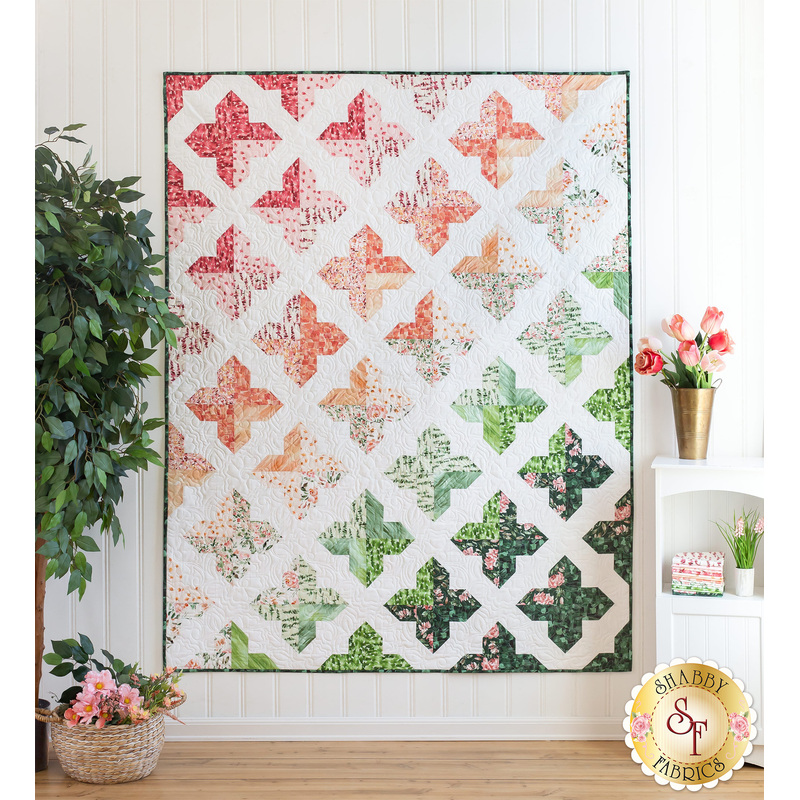 A colorful quilt featuring a geometric pattern of crosses in shades of pink, green, and white hangs on a wall, surrounded by potted plants and decorative items.
