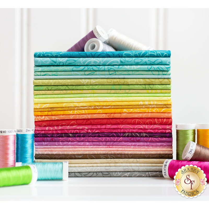 A neatly arranged stack of colorful fabric rolls in various shades, including blues, greens, yellows, oranges, pinks, and purples, sits next to several spools of thread in coordinating colors.