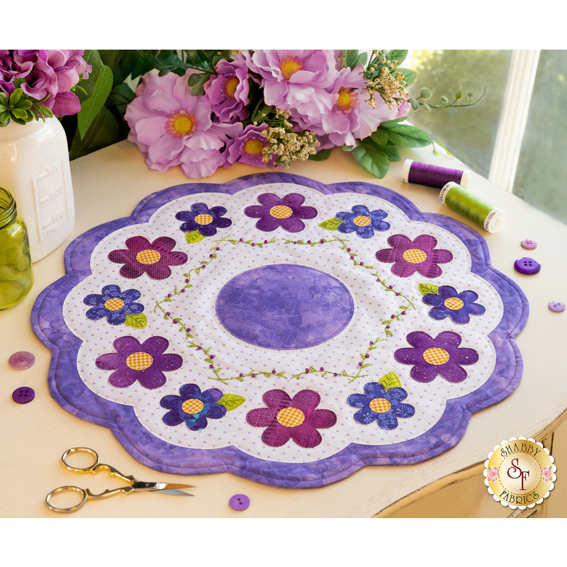 A round floral table mat with a scalloped edge, featuring purple, pink, and blue flowers arranged around a central circle. The mat is placed on a wooden table beside scissors, spools of thread, and decorative buttons, with purple flowers in a vase in the background.