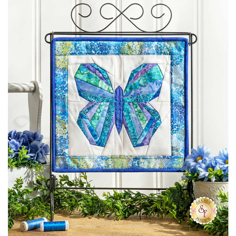 Photo of a blue and green butterfly made with foundation paper piecing techniques, hanging on a craft holder atop a counter with spring decor of flowers, foliage, and matching thread spools