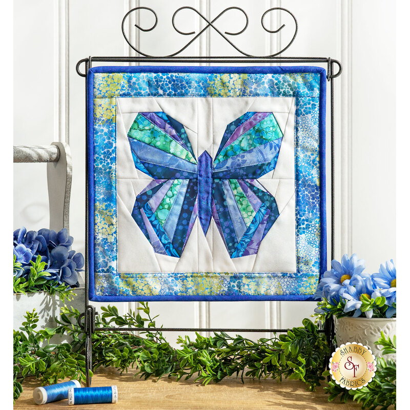 The completed Foundation Paper Piecing Kit - June, a beautiful butterfly colored in blue, teal, and indigo fabrics, displayed on a craft stand and staged with a coordinating décor.