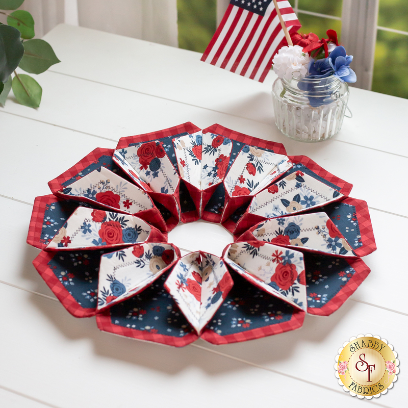 Flower shaped table topper with central opening with a small jar and patriotic decor in the background. Topper is made of crisp white, red and navy blue fabrics with multicolor florals.