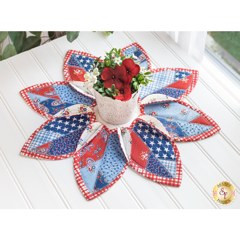 Leaf shaped table topper with central opening containing a floral centerpiece. Topper is made of red white and blue patriotic fabrics.