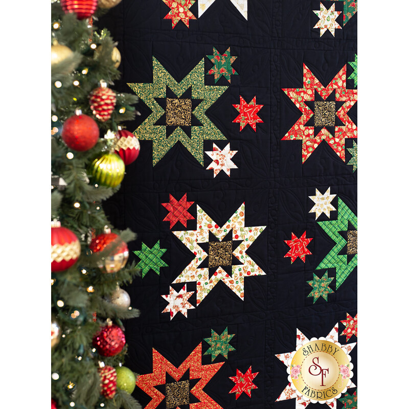 Close up of Merry & Bright Quilt showing a cluster of starbursts made with black, red, green, and cream fabrics with Christmas motifs and metallic accents featuring a decorated Christmas tree in the foreground.