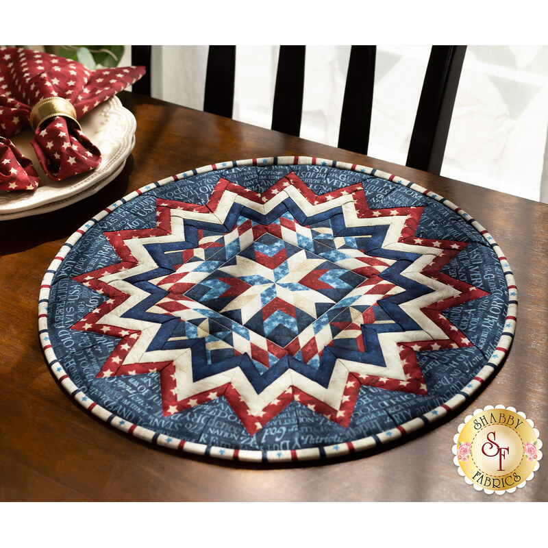 Image of a red, white, and blue circular table topper with a miniature quilt block center piece. The table topper is set on a brown wood table with spotted cloth napkins atop a stack of plates to the side