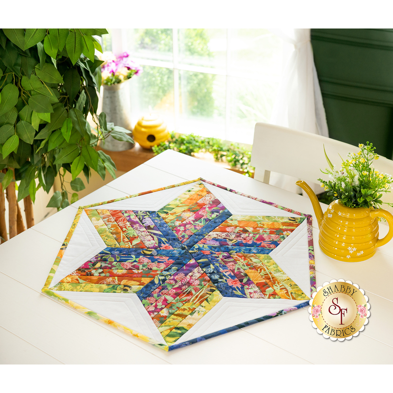 A colorful star-shaped quilt with vibrant floral patterns is displayed on a white table. A yellow watering can and a small potted plant are nearby, with natural light coming through a window in the background.