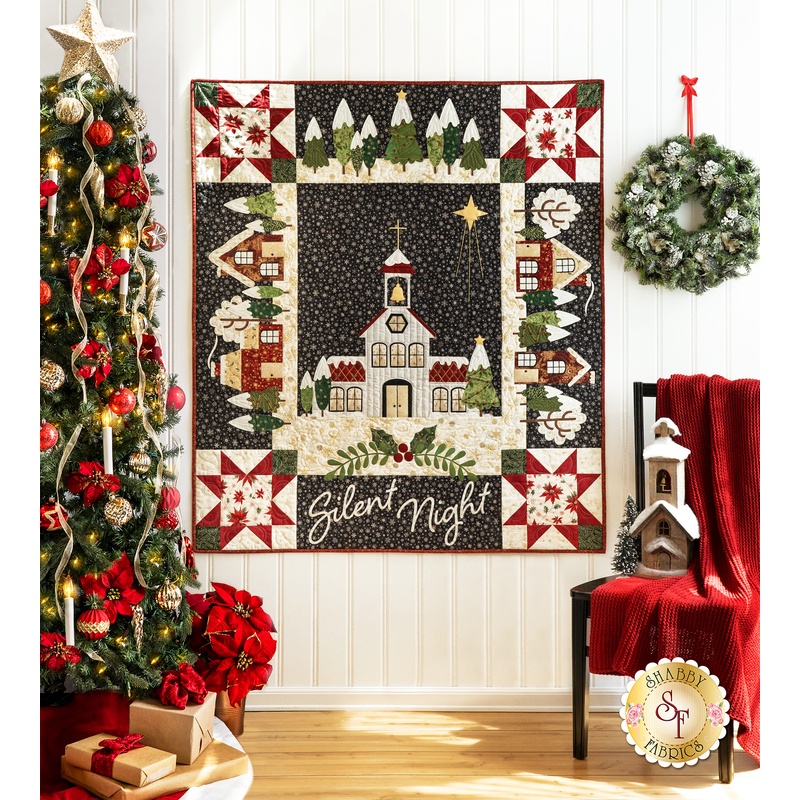 A black, white, and red quilt with a church in the center and village motifs around the border hanging on a white paneled wall with a chair with a red draped blanket on it, and a decorated christmas tree with wrapped gifts on the left