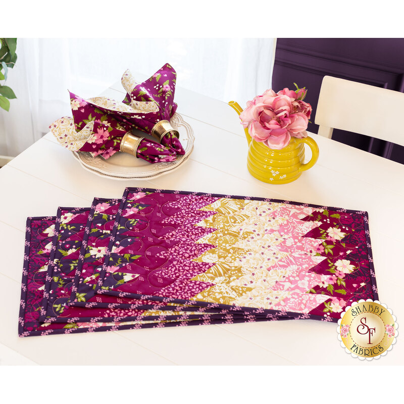 A set of colorful fabric placemats in shades of purple, pink, and gold is laid out on a white table. There is a yellow pitcher filled with pink flowers and matching napkins arranged on a plate nearby.