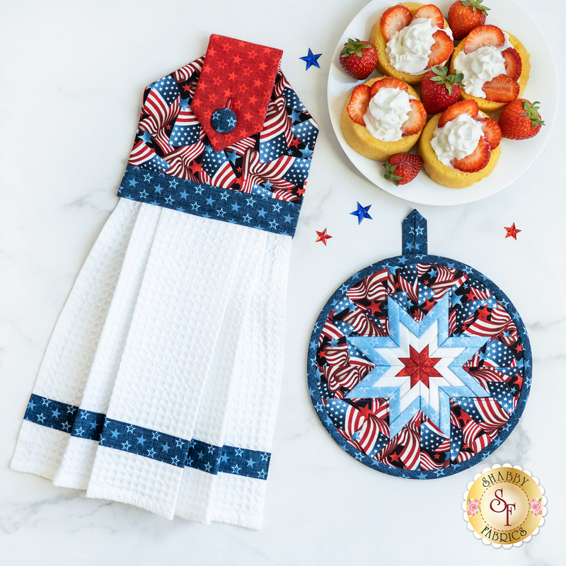 A decorative kitchen towel with a red, white, and blue design hangs next to a round pot holder featuring a star pattern. There is a plate of yellow cupcakes topped with whipped cream and strawberries nearby, along with small star decorations scattered across the surface.