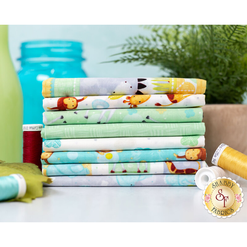 A stack of colorful fabric bundles arranged neatly on a table. The fabrics feature various playful patterns, including animals and playful designs. In the background, there are spools of thread and a green plant, with a blue jar partially visible.