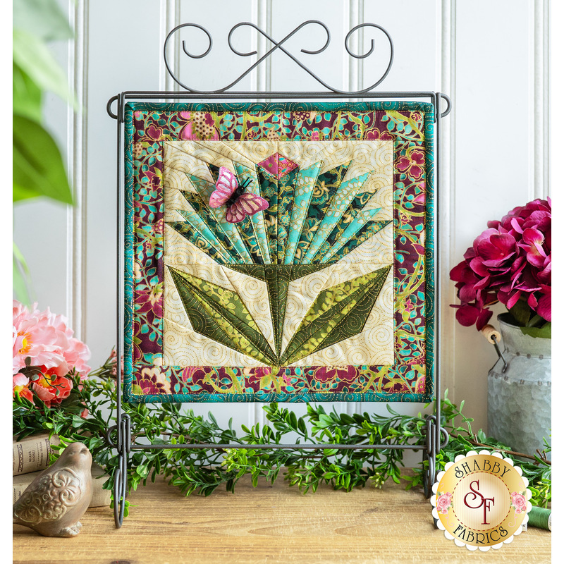 Photo of a completed quilt block featuring a stylized flower with a purple butterfly accent made with metallic fabrics and thread hanging from a craft holder with swirls standing on a countertop against a white paneled wall with floral decorations all around