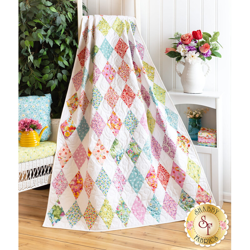 Photo of a multicolored quilt with alternating white diamonds all over, draped in front of a white paneled wall with a white wicker chair, houseplant, and floral decor on one side and a small white shelf with flowers in vases on the other