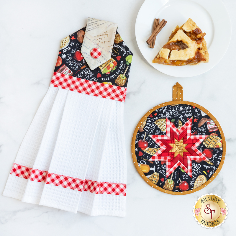 Hanging towel and hot pad for September, featuring pies, fruit, and words with gingham accents on a white countertop with a plate with a slice of apple pie on it