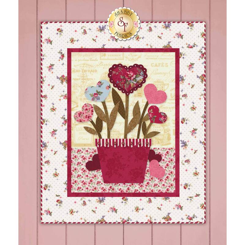 A decorative fabric wall hanging features a pot of flowers with heart-shaped blooms in shades of pink and blue, set against a light floral background and bordered with a red frame.