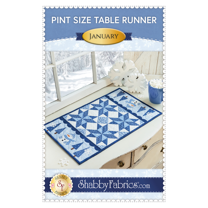 Front cover of the pattern featuring title, designer and photo of finished project--A blue and white winter-themed table runner atop a white counter with a wintry scene outside an adjacent window and snowflake decorations with a mug of hot cocoa