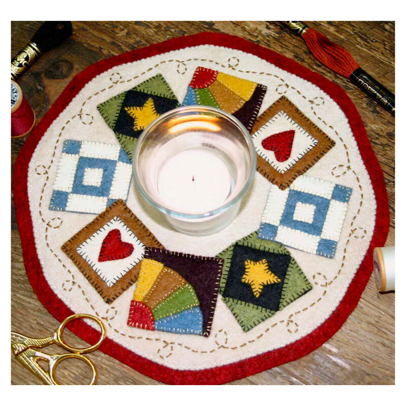 A round decorative placemat featuring colorful quilt-like patches in various shapes, with a glass candle placed in the center. Surrounding materials include sewing scissors and spools of thread.