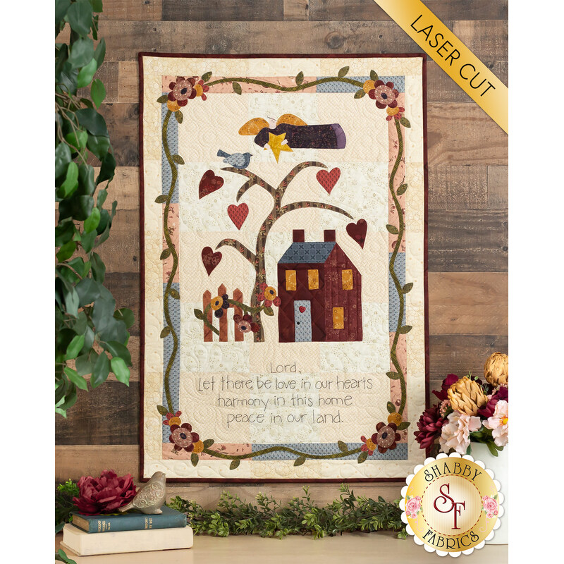 A quilted wall hanging featuring a tree with hearts, a house, and a fence. The design includes a bird, flowers, and text that reads, Lord, let there be love in our hearts, harmony in this home, peace in our land. The quilt is framed by decorative borders and has a label in the corner that says Shabby Fabrics.