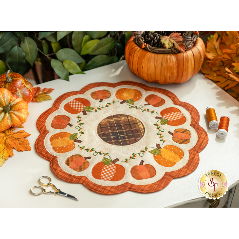 Photo of a scalloped table mat with pumpkins in a ring and embroidered details on a white table with a pair of scissors, threads, and pumpkin decor