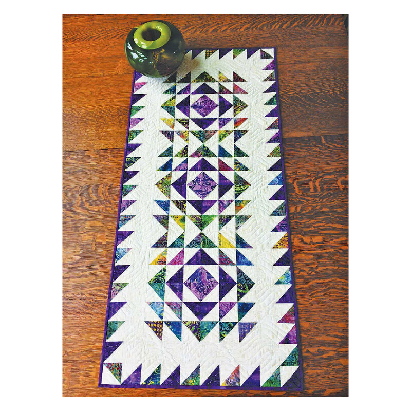 Photograph of the finished vortex table runner, including a white background with purple and green triangles on it, set on a wooden table 