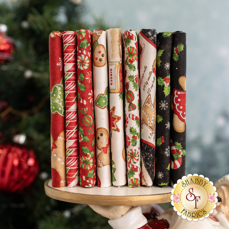 A stack of patterned fabric rolls arranged vertically on a round wooden stand. The fabrics feature various holiday-themed designs, including gingerbread cookies, candy canes, and snowflakes, in festive colors like red, green, and white. In the background, there is a blurred festive setting with Christmas decorations.