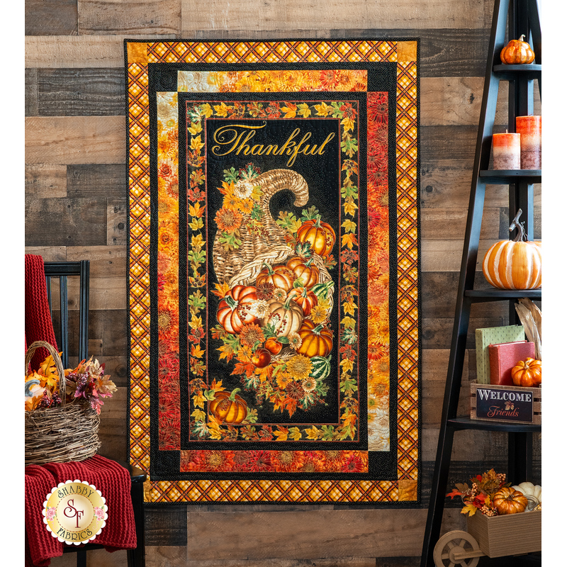 Photo of a Thanksgiving and autumn-themed panel quilt hanging on a brown paneled wall with fall decor like pumpkins, candles, and books on a shelf with a floral basket on a chair in the foreground