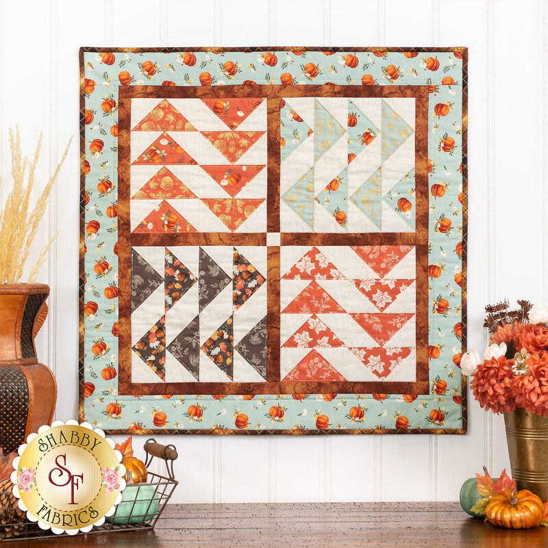 Photo of a wall hanging designed in quadrants of flying geese units made with aqua, orange, cream, and brown autumn themed fabrics hanging on a white paneled wall with fall decor on the countertop below it