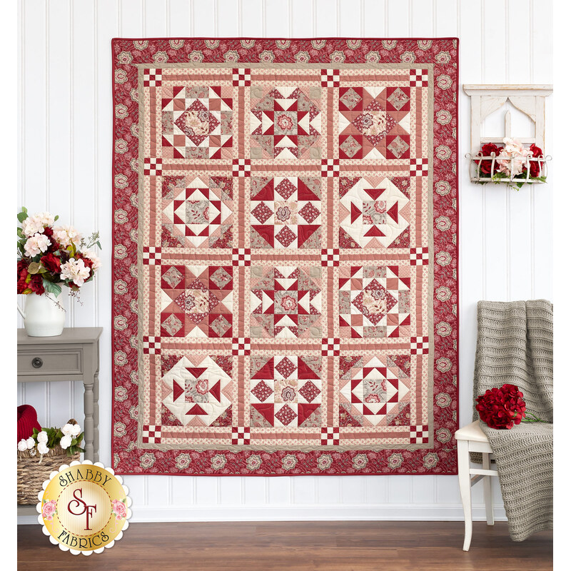 Photo of a large red and cream patchwork quilt hanging on a white paneled wall with taupe and white furniture and red flowers all around.