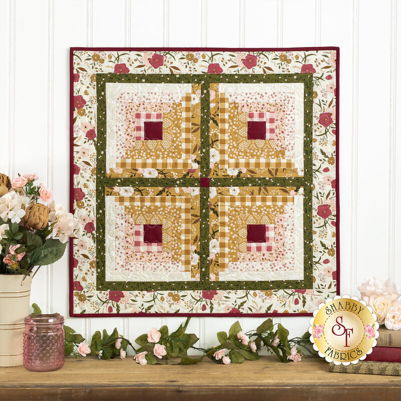 Photo of a floral wall hanging on a white paneled wall with floral decor, books, and a pink jar on a brown countertop at the bottom of the image