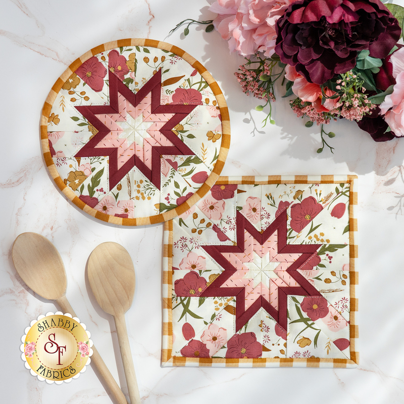 Photo of two folded star hot pads, one round and one square, made with pink, white, and red floral fabrics and yellow gingham laying flat on a white countertop with wooden spoons and a bouquet of flowers