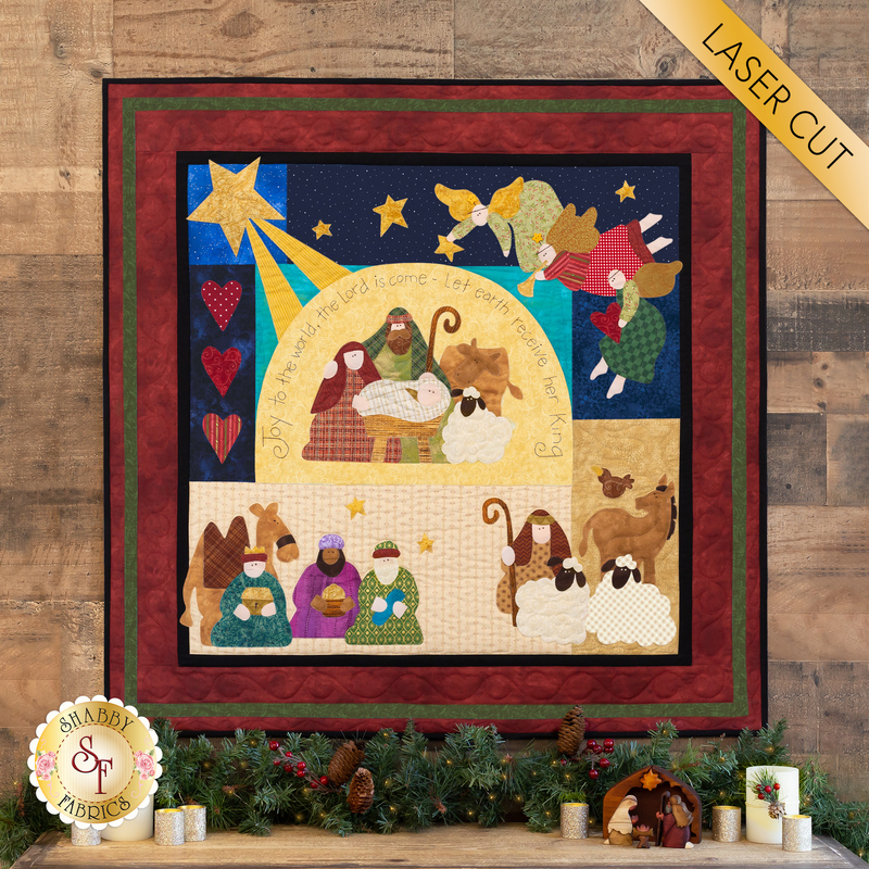 A colorful quilt design featuring a nativity scene with Mary, Joseph, and baby Jesus surrounded by animals. The quilt includes stars, hearts, and angels, with a border of red fabric. A label in the top right corner reads LASER CUT. The background appears to be wooden, with festive decorations at the bottom, including greenery.