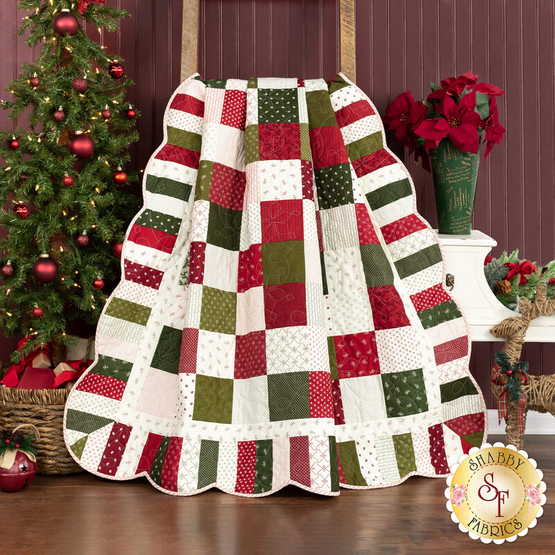 Photo of a green, red, and cream Christmas-themed quilt draped over a ladder in front of a red wall with a Christmas tree and poinsettia decor in the background.