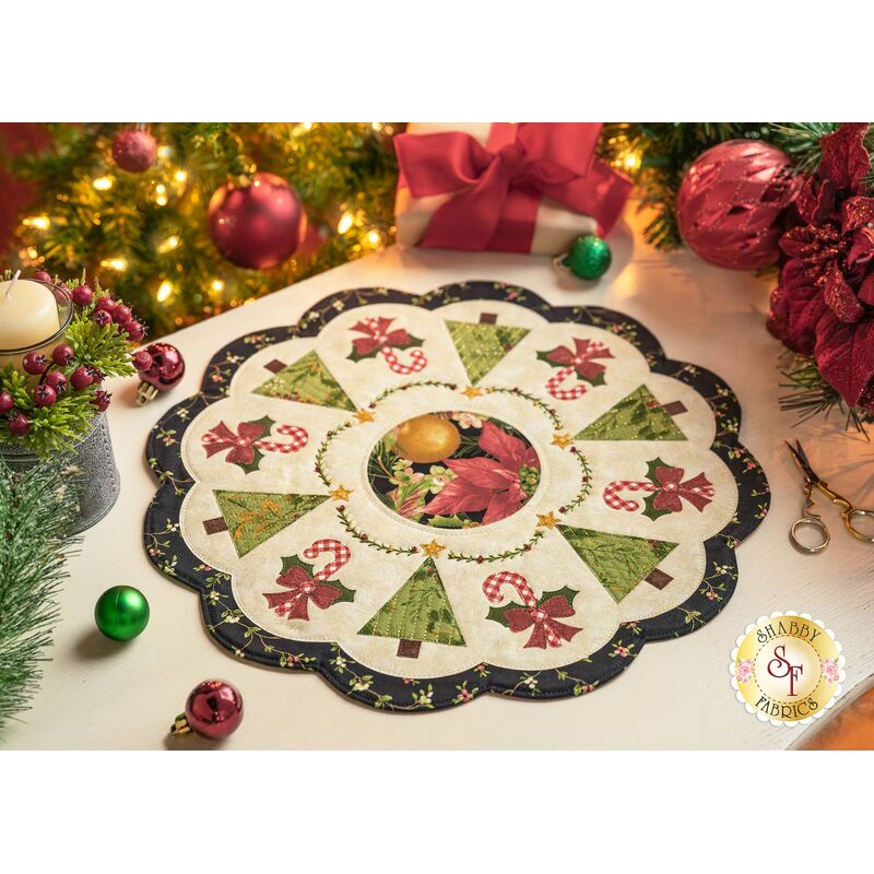 A decorative holiday table centerpiece with a floral and Christmas tree design, featuring green trees, red poinsettias, and candy canes, set on a light background with a scalloped edge. Surrounding the centerpiece are festive decorations including a lit candle and ornaments.