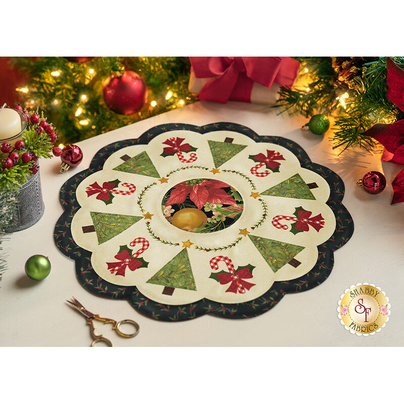 A decorative holiday table centerpiece with a floral and Christmas tree design, featuring green trees, red poinsettias, and candy canes, set on a light background with a scalloped edge. Surrounding the centerpiece are festive decorations including a lit candle and ornaments.