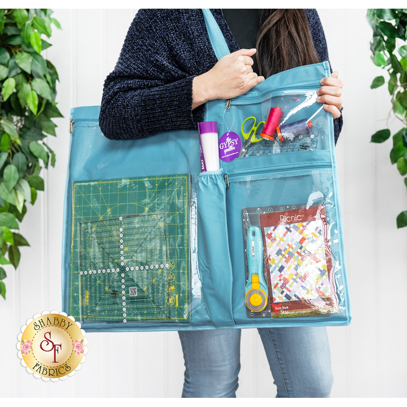 A person holding a turquoise craft tote filled with sewing tools, fabric, and a cutting mat, surrounded by greenery.