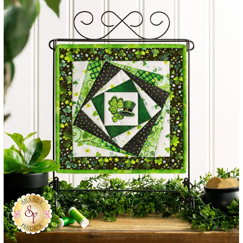 Photo of the finished wall hanging featuring offset squares in shades of green with a shamrock and top hat in the center, on a craft holder in front of a white paneled wall and decorative foliage.
