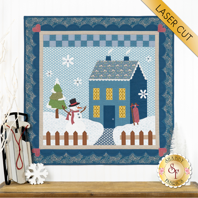 The completed Little Garden House in Winter wall hanging in a soft winter palette of navy, blue, and white, hung on a white paneled wall and staged with coordinating snowy decor.