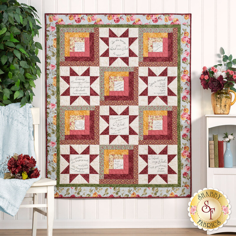 Photo of a red, cream, pale blue, and golden yellow quilt made with traditional quilt star piecing designs and rectangular blocks with quotes from psalms interspersed, hanging on a white paneled wall with houseplants, red flowers, and decor on either side.