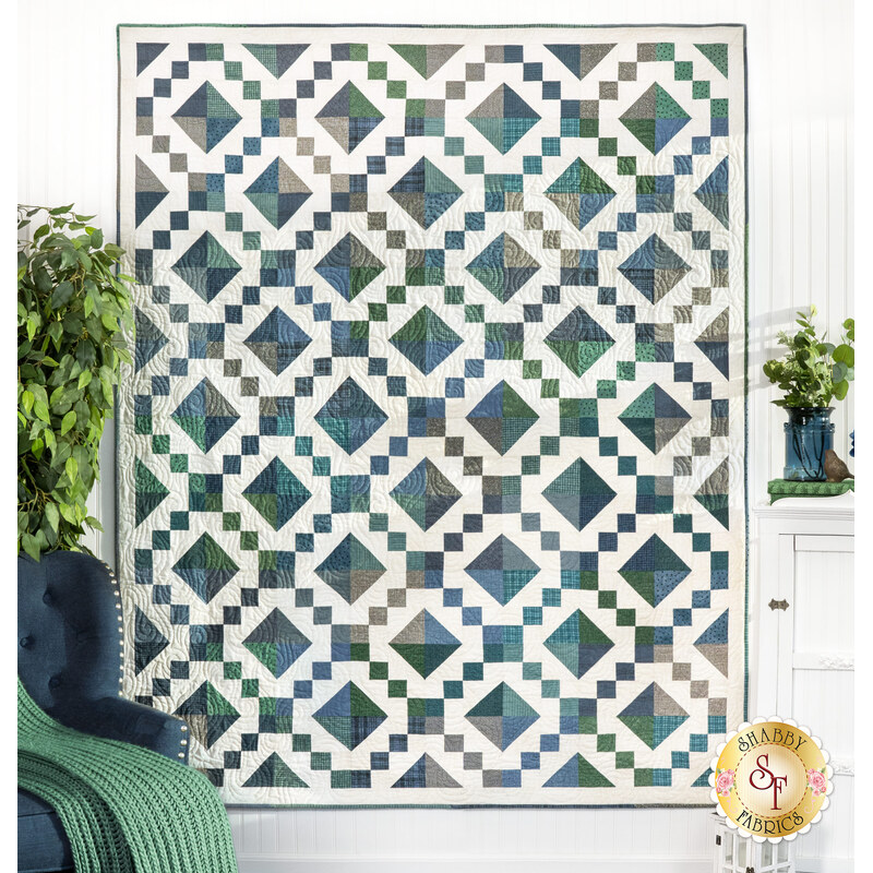 The completed Sunset Road Quilt, colored in cream, blue, teal, and green fabrics from the Woolies Flannels collection by Maywood Studio, hung on a white paneled wall and staged with complementary decor and furniture.